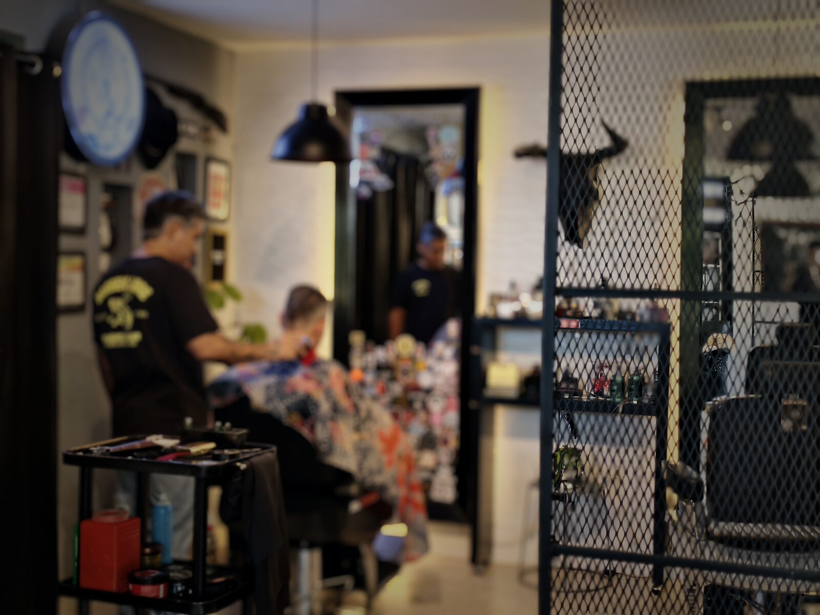Un barbero corta el pelo a un cliente en Barbero Libre; al fondo, una mampara de malla metálica y un círculo de neón azul. Las personas aparecen desenfocadas.