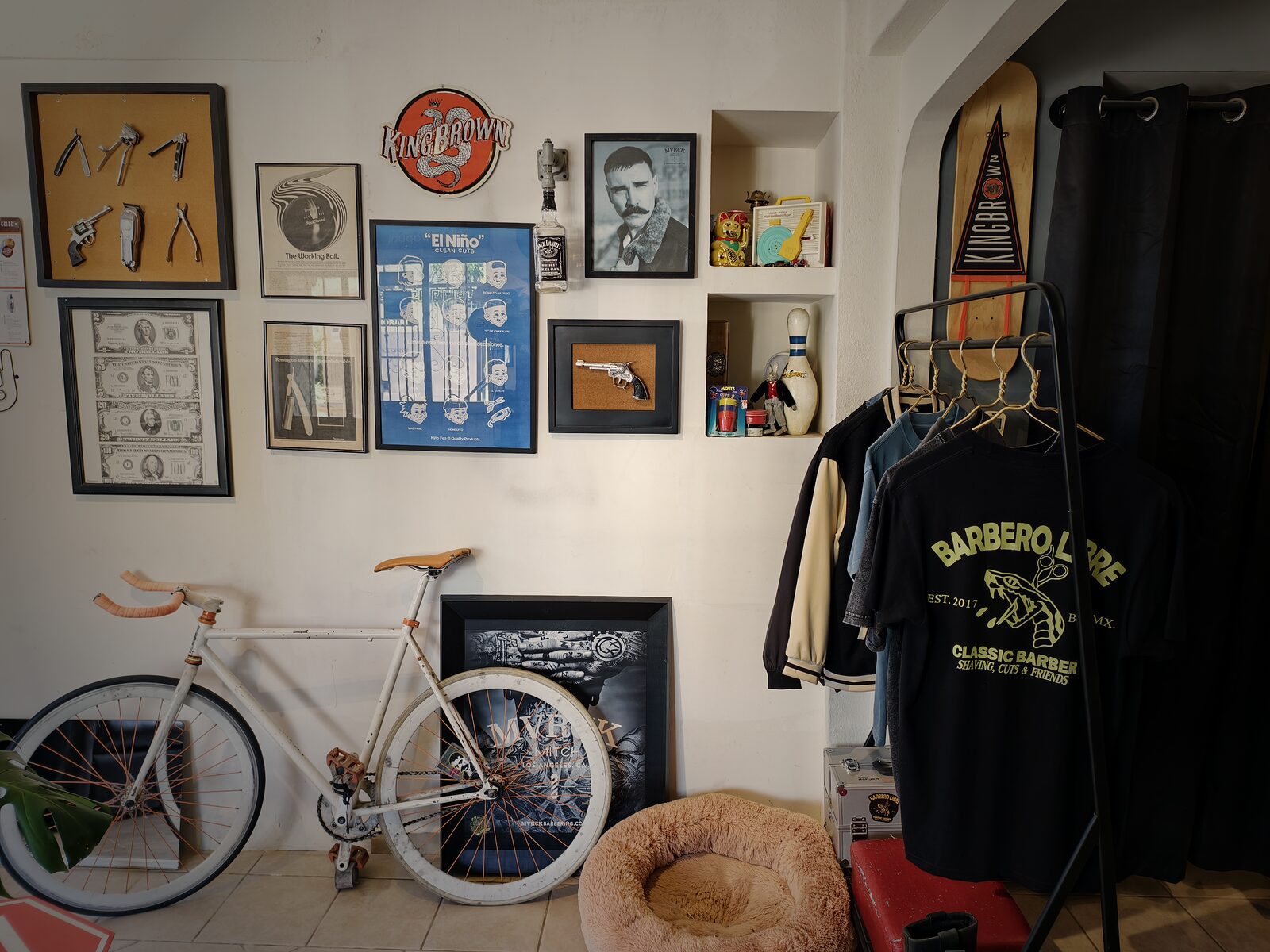 Pared con navajas y máquinas enmarcadas, billetes antiguos, el póster “El Niño clean cuts” y un letrero de KingBrown; abajo, una bicicleta fixie blanca y un perchero con una playera de Barbero Libre.