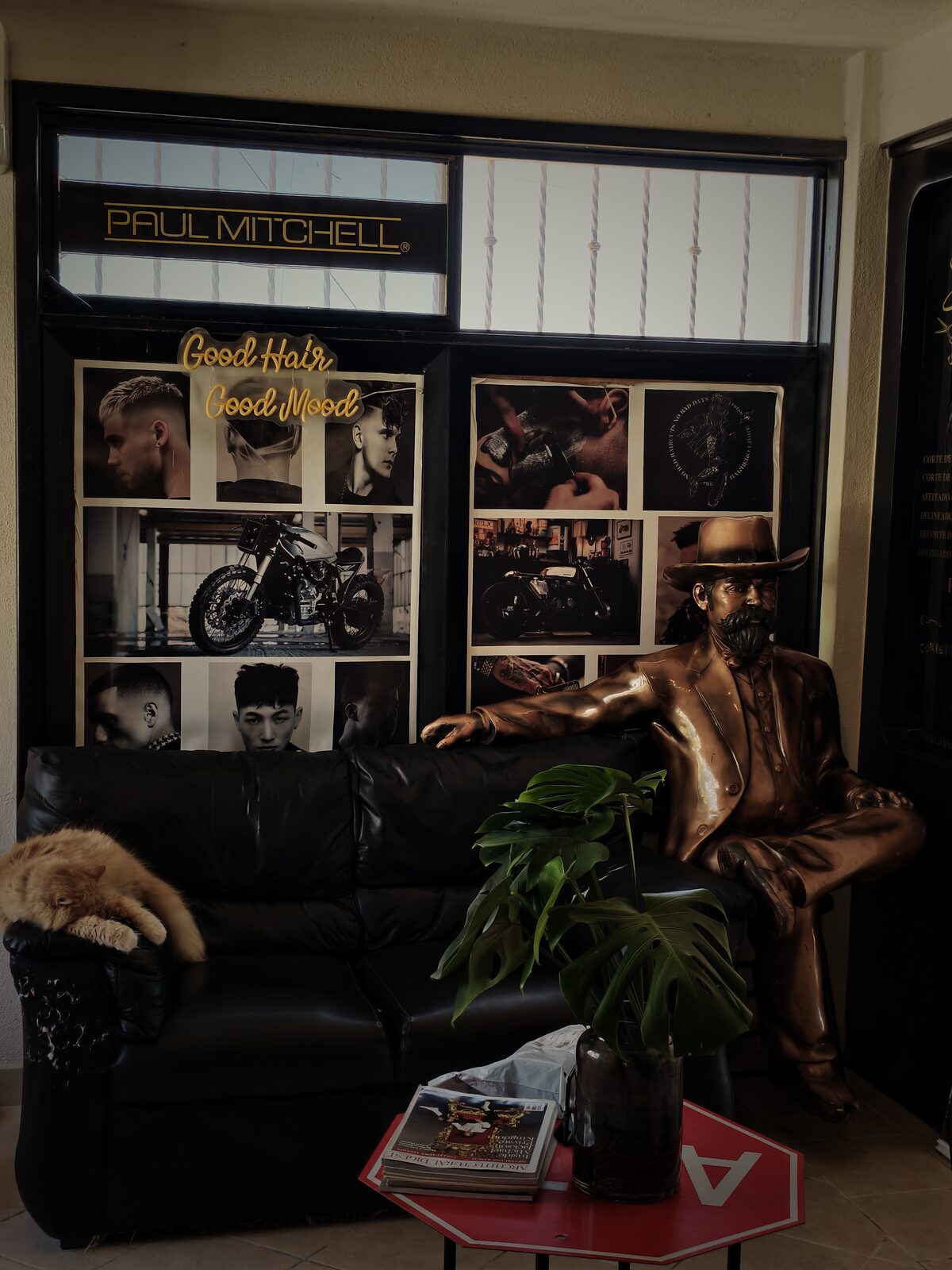 Sala de espera con un sillón de piel negra, un gato naranja dormido, una estatua de bronce de un vaquero y un collage con motos café racer bajo el neón “Good Hair Good Mood”.
