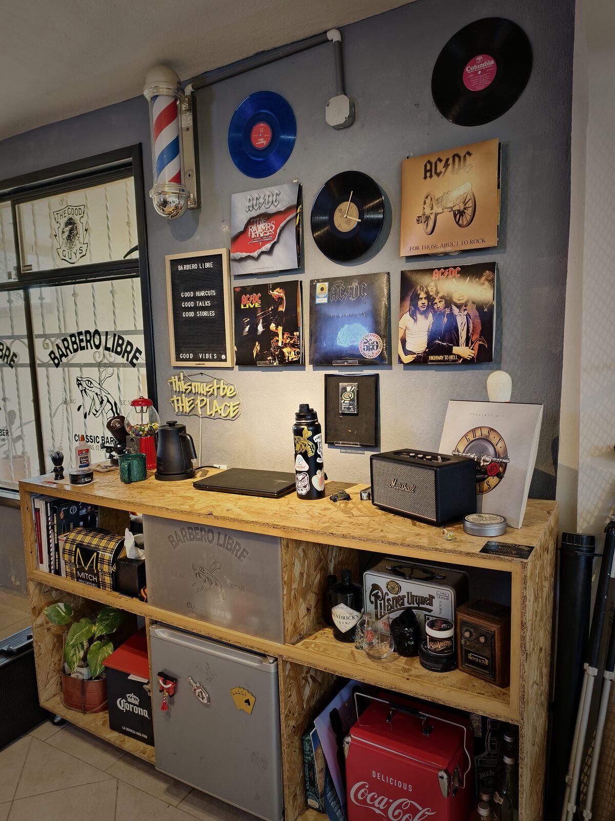 Pared de vinilos con discos de AC/DC y Guns N’ Roses, un poste de barbero y una bocina Marshall; un letrero dice “Good haircuts, good talks, good stories, good vibes” y un neón, “This must be the place”.